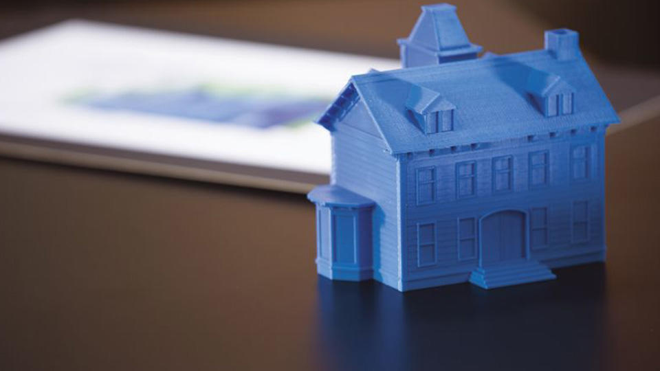 3D printed blue house on desk next to blurred document