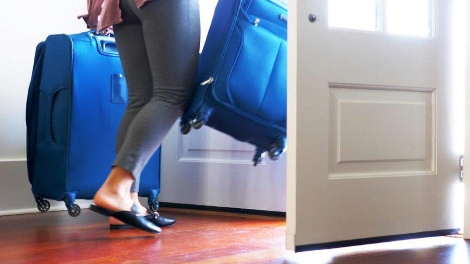 A traveler strides out her front door with 2 rolling luggage cases