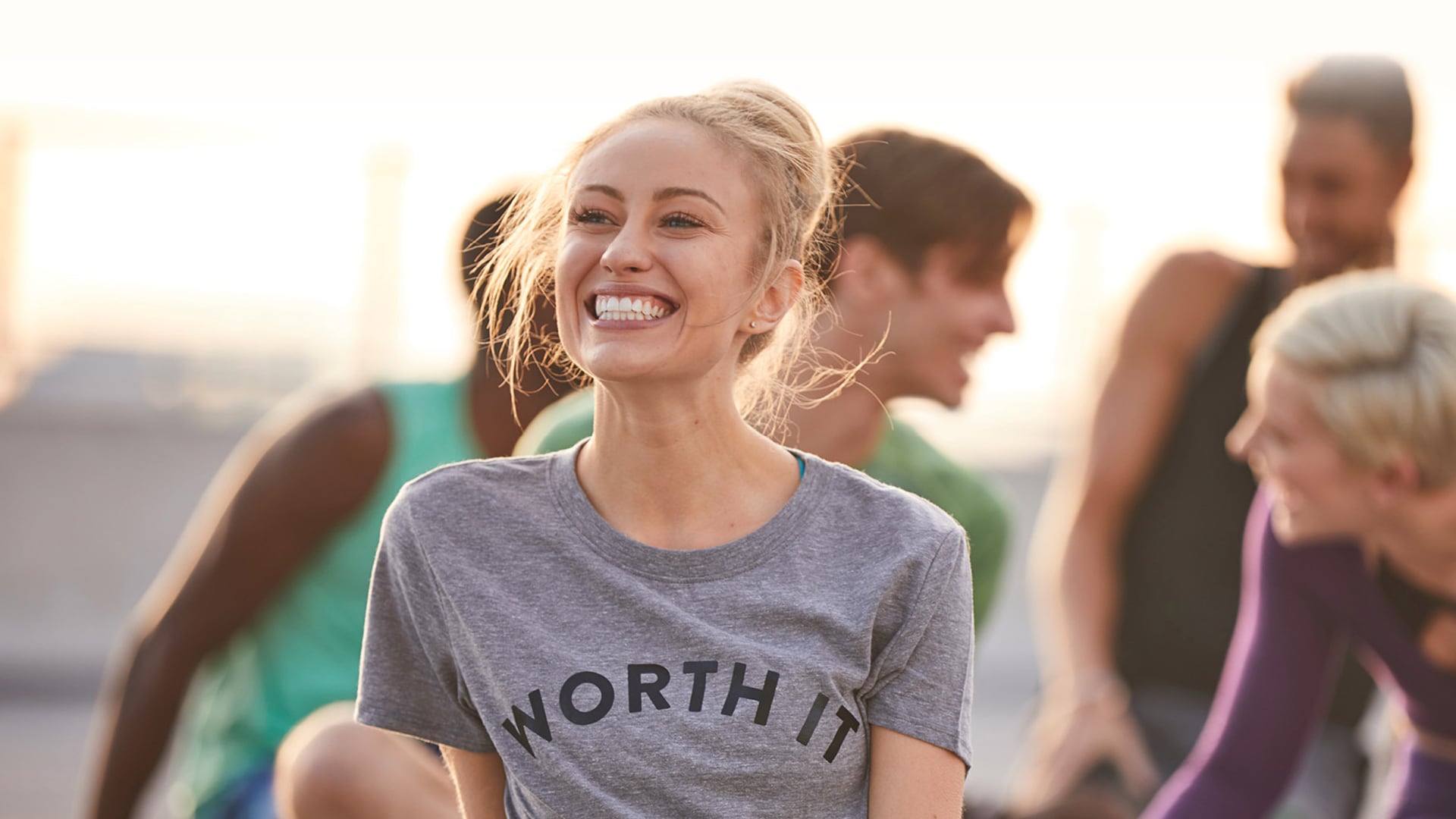 Une jeune femme portant un t-shirt gris avec écrit « Worth it » sourit tandis qu'un groupe de personnes en pleine conversation sourit et rit derrière elle.