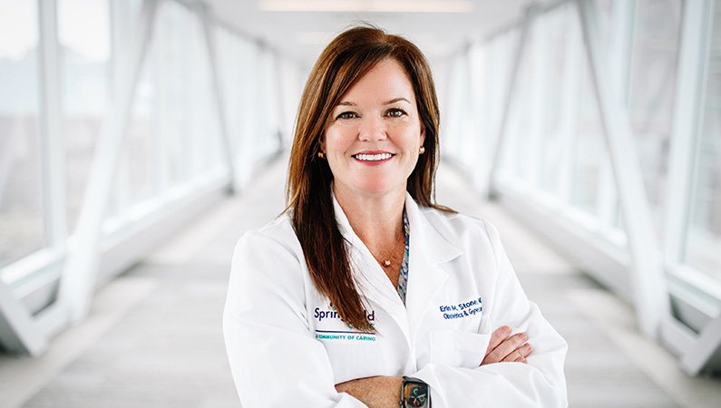 Female in white coat smiling in a naturally lit hallway.