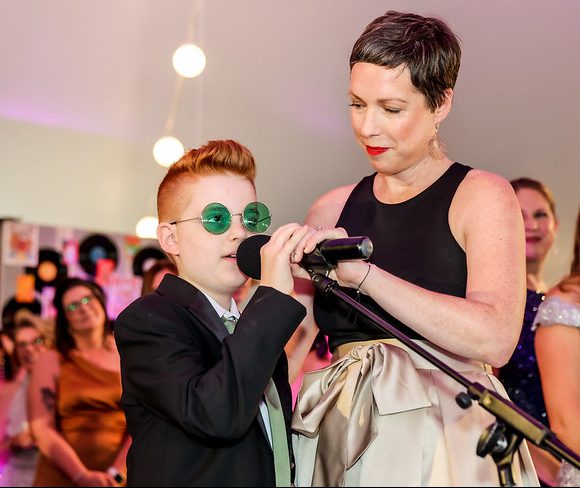 A mother and son in formal attire stand on stage to speak at a fundraising event