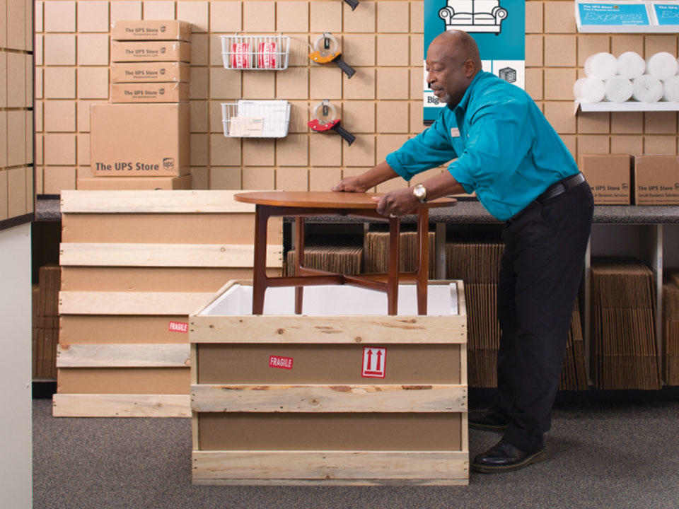 Male The UPS Store associate placing a wooden coffee table into freight container in the store