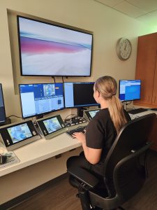 Female medical provider in black scrubs and a long ponytail sits at computer console with multiple screens.