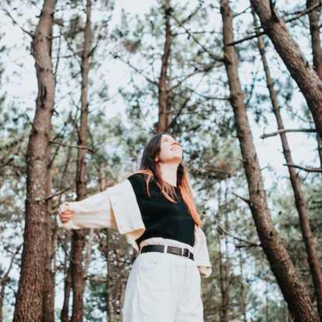A person stands in a serene forest, arms outstretched, eyes closed, smiling. Tall trees surround, creating a peaceful, uplifting atmosphere.