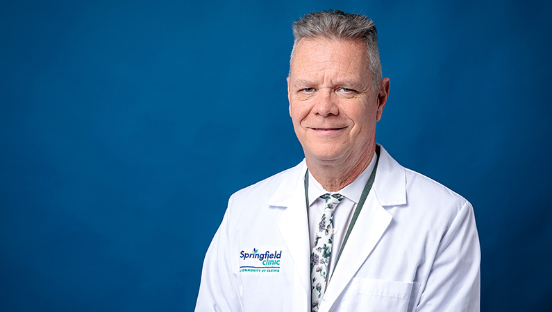 Male neurology doctor in white lab coat smiling in front of navy blue backdrop.