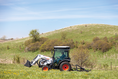 Bobcat construction tractor with three-point finish mower driving through hilly countryside.