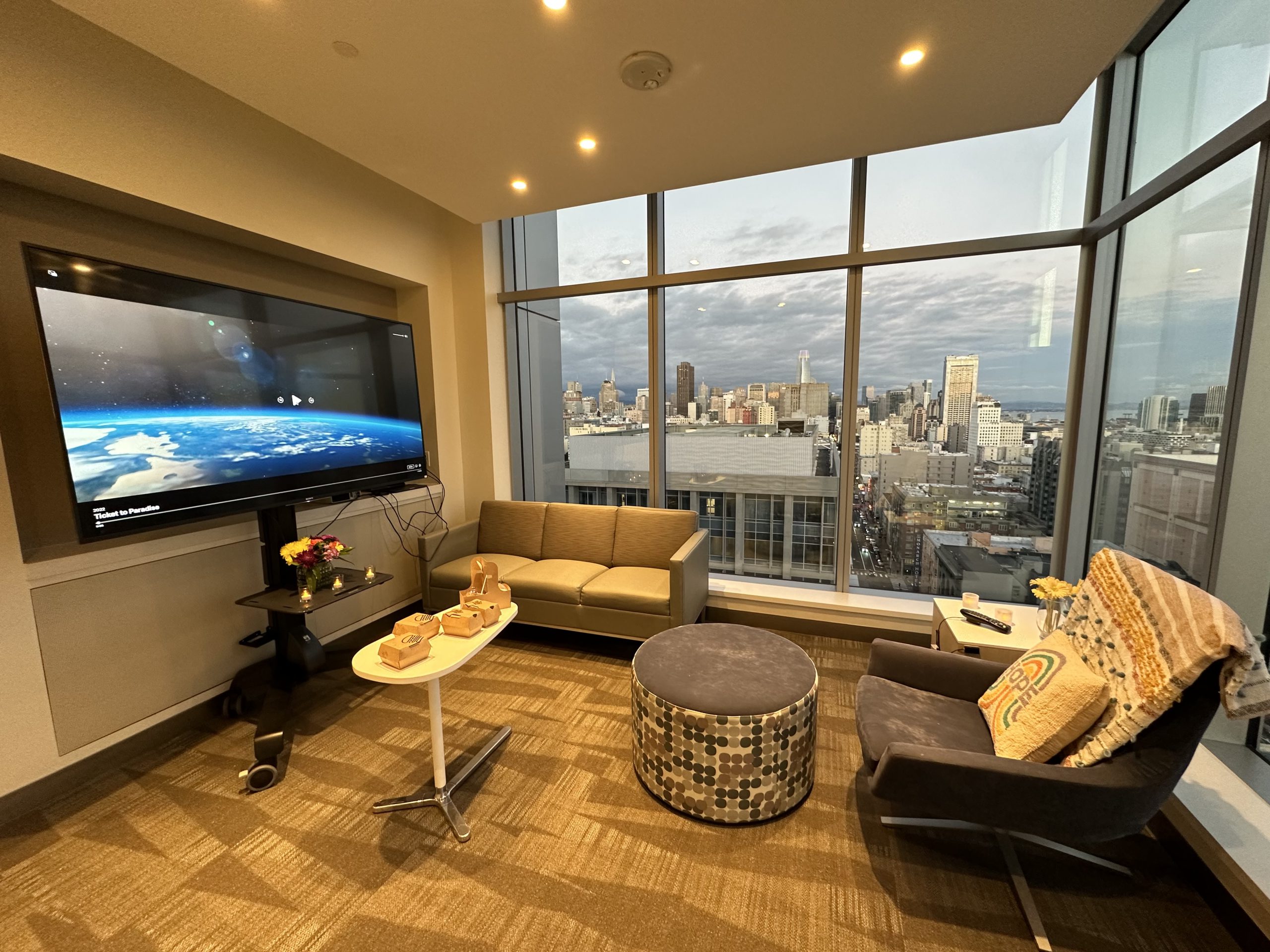 A hospital lounge with floor to ceiling windows is set up for a date night with takeout food, flameless candles and movie screen, and cozy throws and pillows.