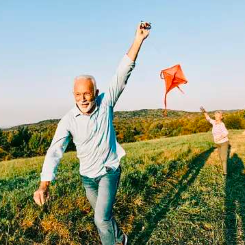 Elderly man joyfully flies a red kite in a sunny grassy field with rolling hills in the background, conveying happiness and freedom.
