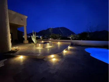 Troon North backyard at night showing the full travertine patio, fire pit, pool, and spa with Pinnacle Peak in the background. LED step lighting across the tiered deck keeps the space usable after dark. This is what a complete Scottsdale backyard remodel looks like when everything is designed together. Built by Hacienda Outdoors, ROC #325952.