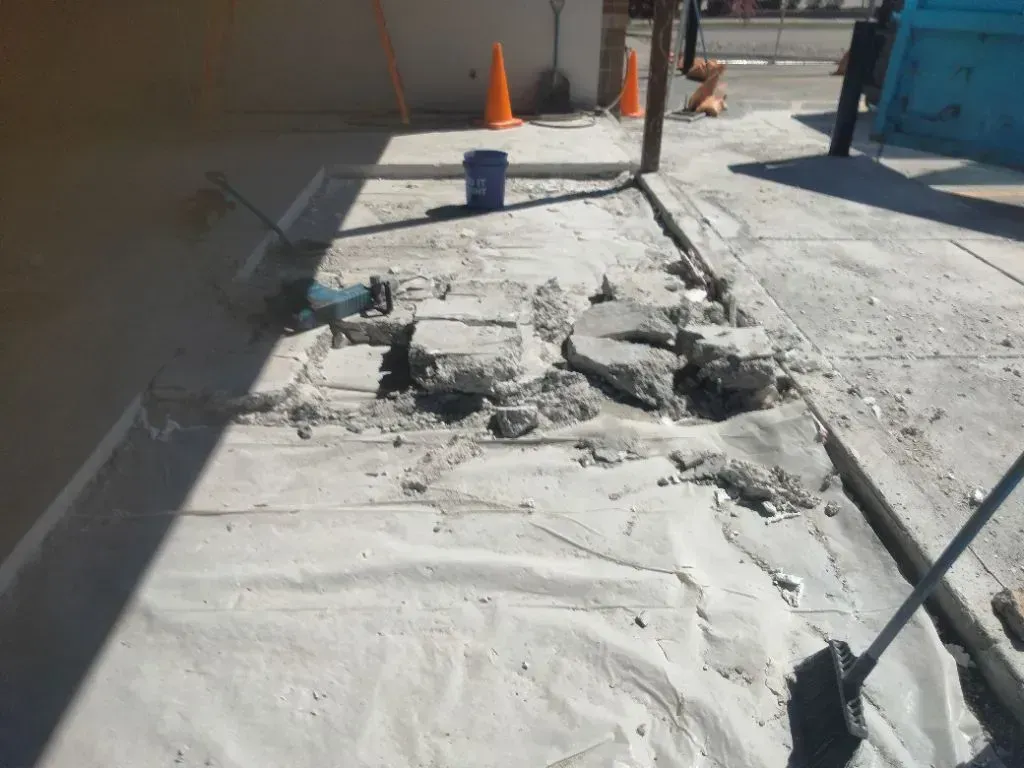 Sidewalk demolition in progress with broken concrete slabs, debris, and tools scattered across work  