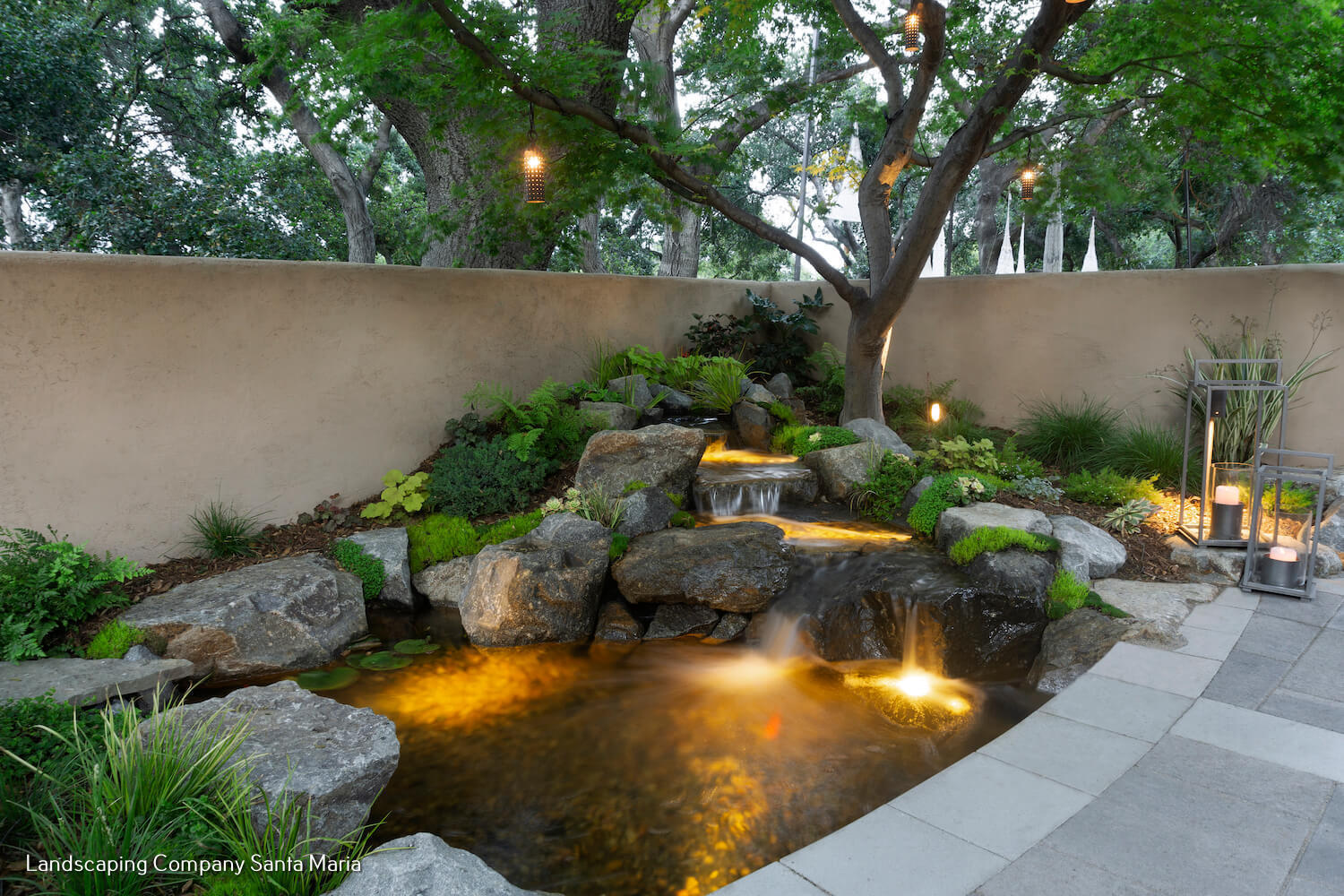 Tranquil landscaped garden pond with cascading rock waterfall, glowing underwater lights, lush greenery, trees, and a tiled patio at dusk.