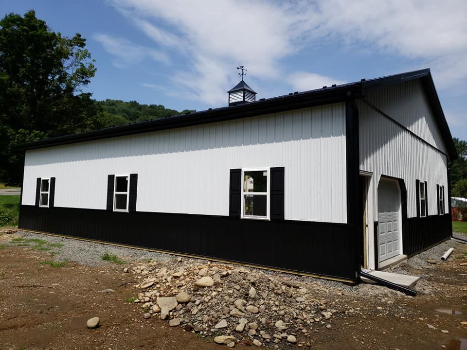 A long metal building with white upper siding and black lower panels, featuring multiple windows with shutters, a garage door, and installed gutter system, set on a rocky, unfinished surface.