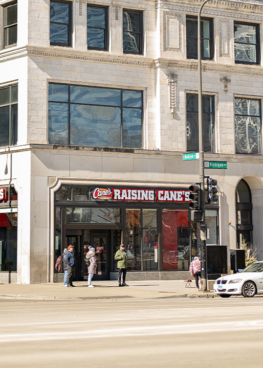 The exterior storefront of a Raising Cane's located on the ground floor of a large stone building on a sunny day, with street signs visible for "Madison" and "Michigan Ave" near a busy intersection.