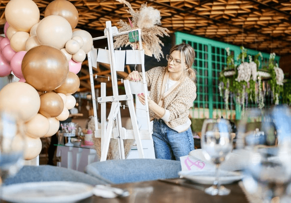 A woman in a cardigan and jeans is arranging small white cards on an easel. She is in a room that appears to be set up for an event, with balloons and table settings visible. In the background, there is a green-paneled wall and hanging floral decorations.