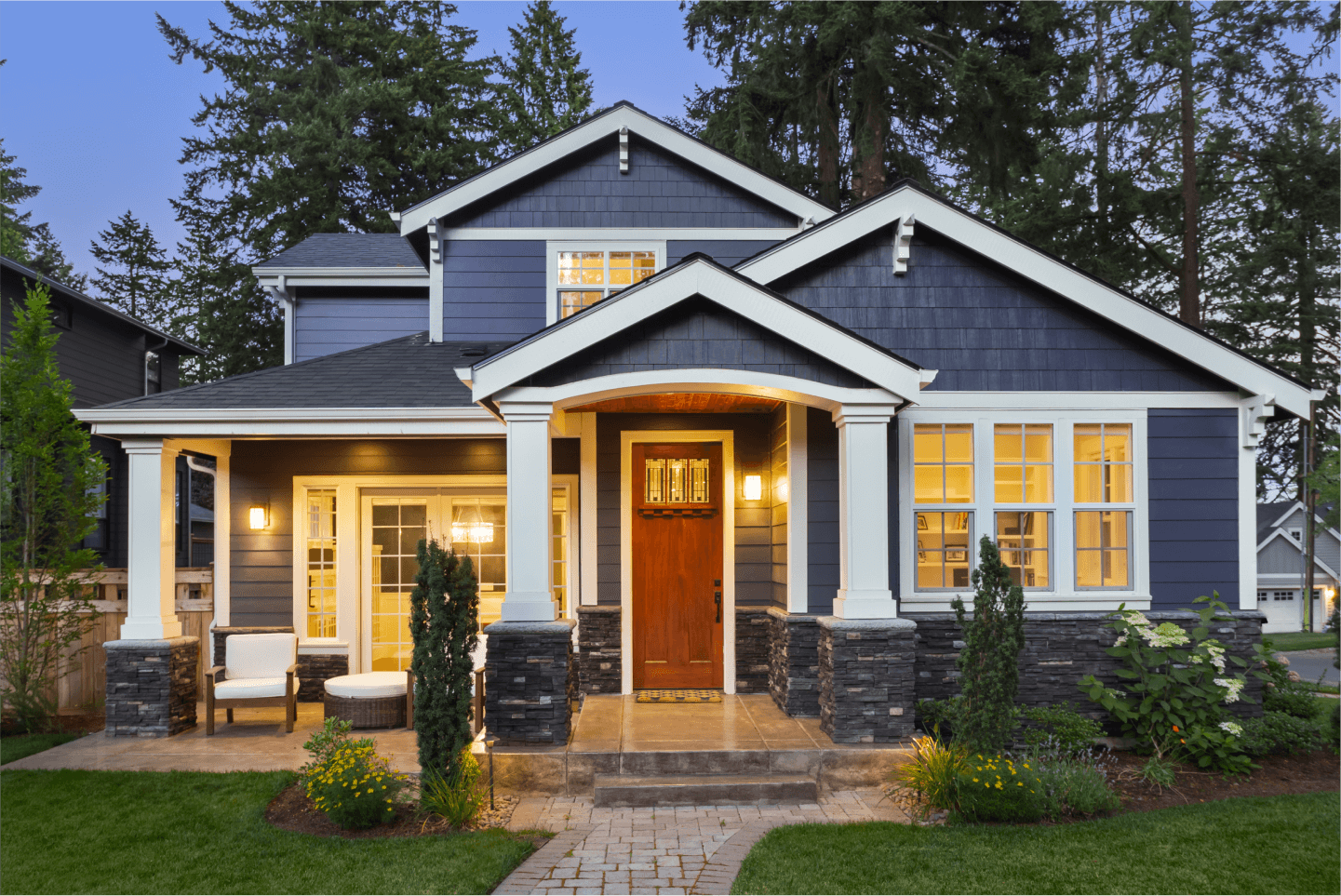 A beautiful, two-story dark blue home with white trim and a warm wood front door is illuminated by interior lights and exterior lamps at twilight, surrounded by lush green grass, manicured bushes, and tall evergreen trees.