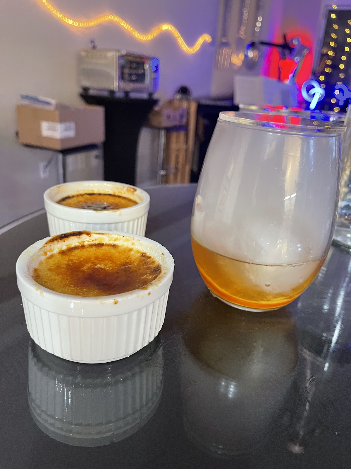 A close-up shot features two ramekins filled with crème brûlée, each with a perfectly caramelized sugar top, sitting on a reflective dark surface. To the right, a stemmed glass holds a amber-colored beverage, possibly whiskey or brandy, filled with ice and swirling with white smoke, suggesting it's a smoked cocktail or a drink served under a smoke-filled cloche. The background is dimly lit with blurred colorful lights and what appears to be a kitchen or bar area with some equipment like a small oven and boxes.
