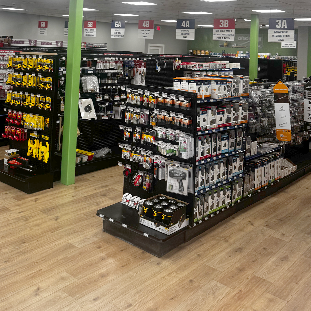 Wide view of the Builders FirstSource hardware area near the entrance, with black shelving filled with locksets, door hardware, batteries, measuring tools and other supplies. A central display holds packaged hardware and bright yellow tie-down straps. Overhead signs identify sections for locks, fasteners, saw blades, drywall tools and accessories, adhesives, and sandpaper.