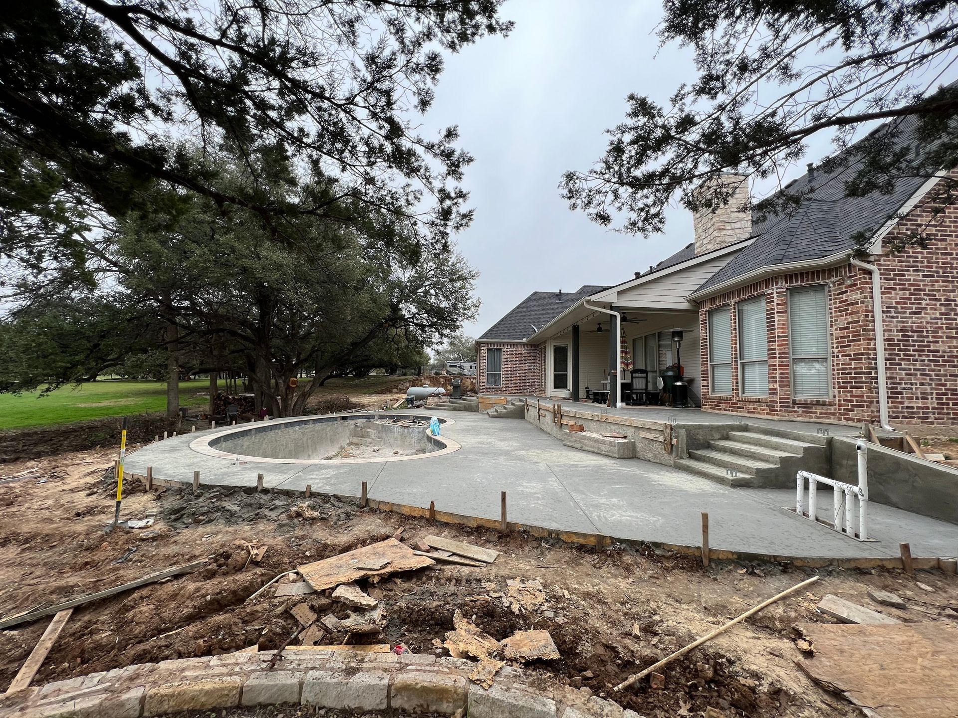 This wide-angle view shows a multi-level concrete pool deck and patio extension under construction behind a large brick home. The curved pool shell is set within the new expansive concrete surface.