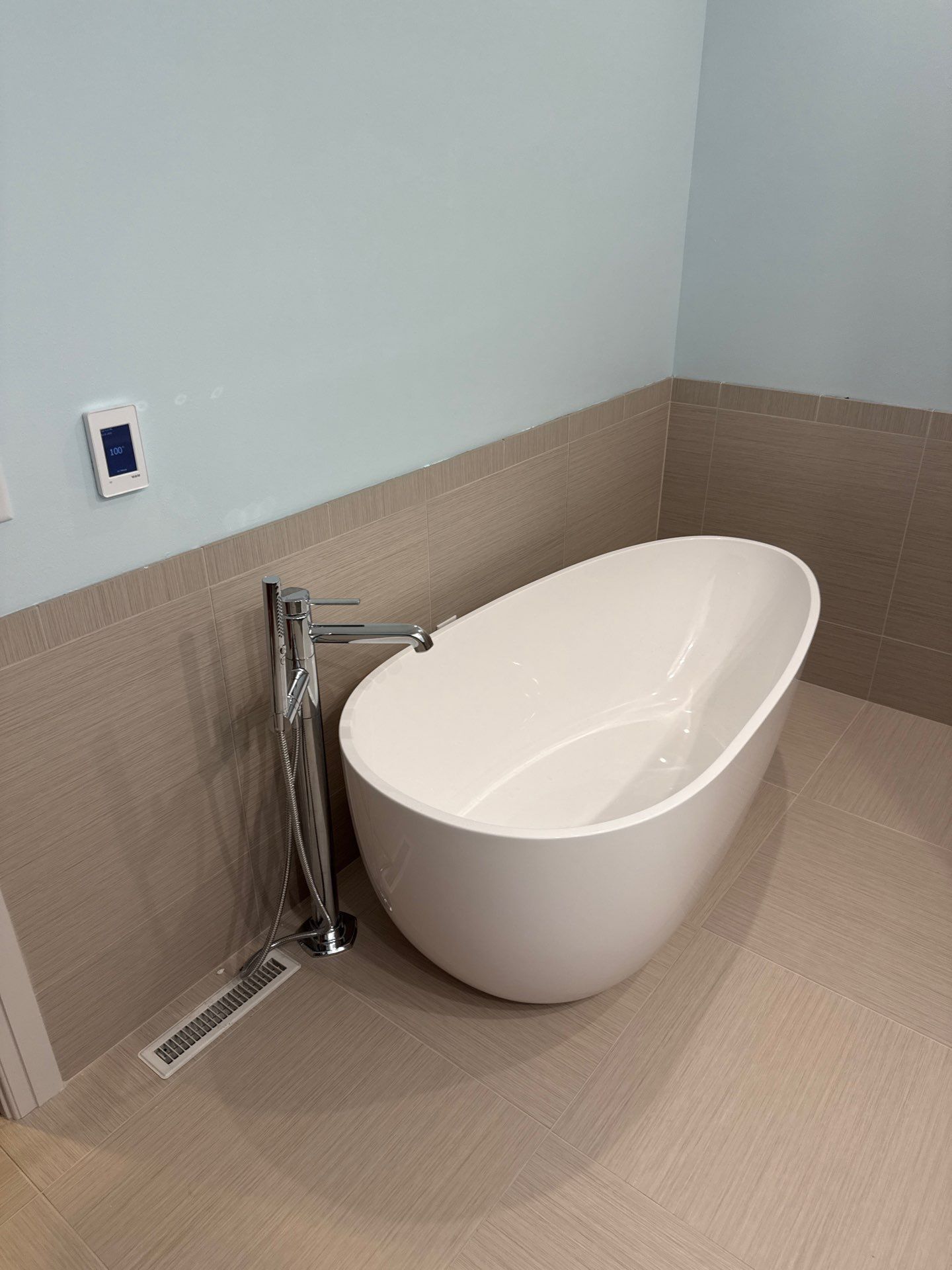 Freestanding white oval bathtub sits in a tiled bathroom corner beside a chrome floor-mounted filler, handheld sprayer, linear floor drain, beige wall tiles, light blue walls, and digital control panel.