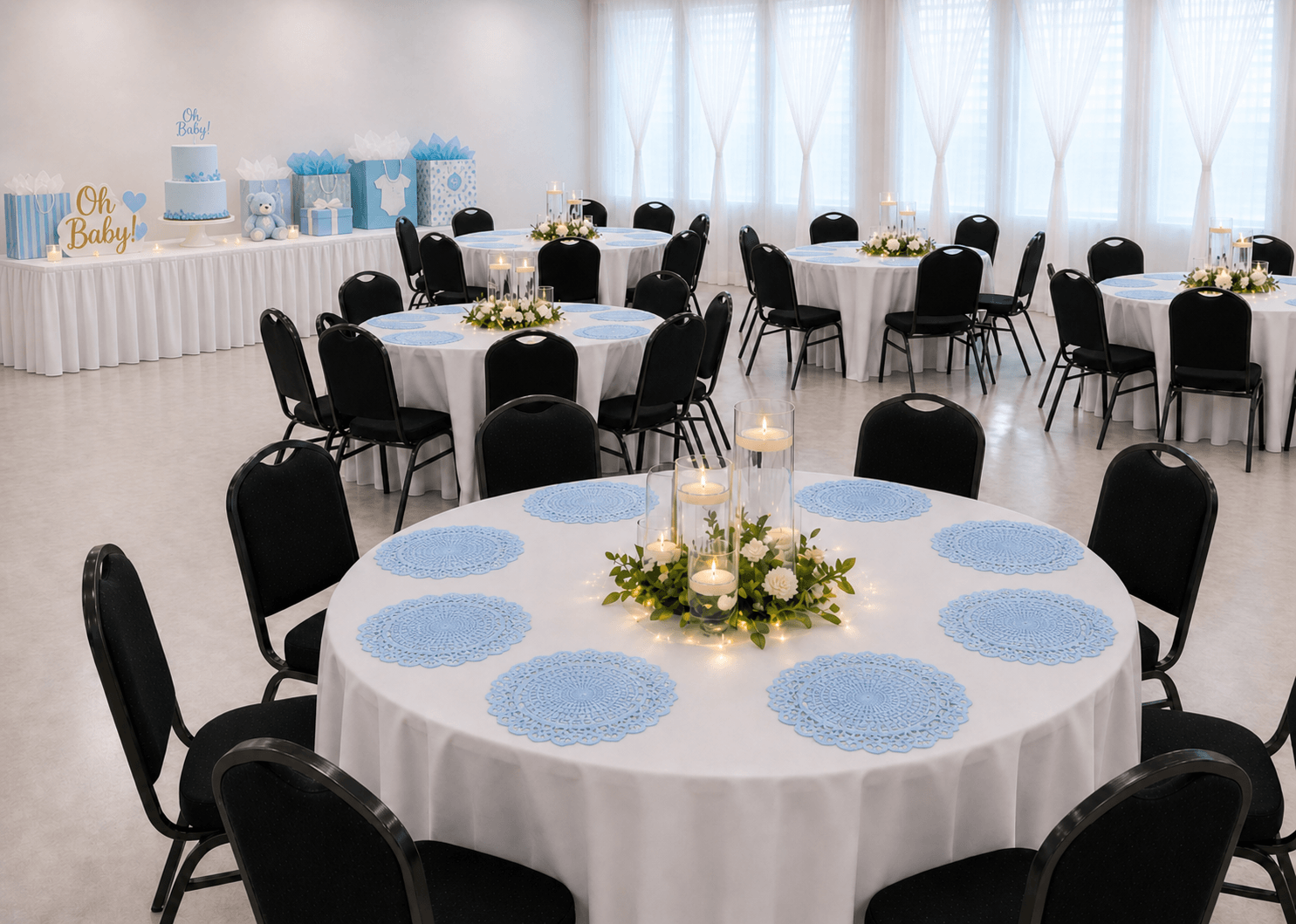 A baby shower setup with round tables covered in white tablecloths, each adorned with blue doilies and floral centerpieces with candles. A dessert table in the background features a light blue tiered cake with 'Oh Baby!' toppers, along with decorative gift bags and a teddy bear.