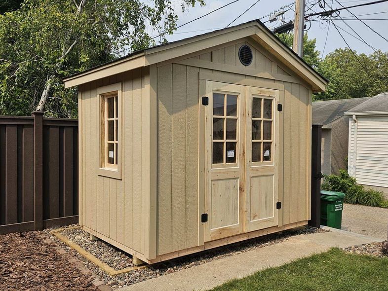 A newly installed tan-colored storage shed with a gable roof, featuring a single window and double french doors with glass panes. The shed sits on a small bed of mulch and gravel next to a concrete walkway in a backyard.