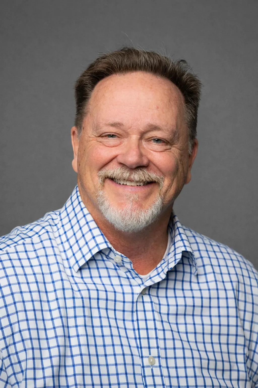 A medium shot portrait of a middle-aged man with a salt-and-pepper beard and mustache, wearing a blue and white checkered button-down shirt. He has light blue eyes and is smiling broadly, looking directly at the camera. The background is a solid, dark gray color. The lighting is soft and even, suggesting a professional headshot.