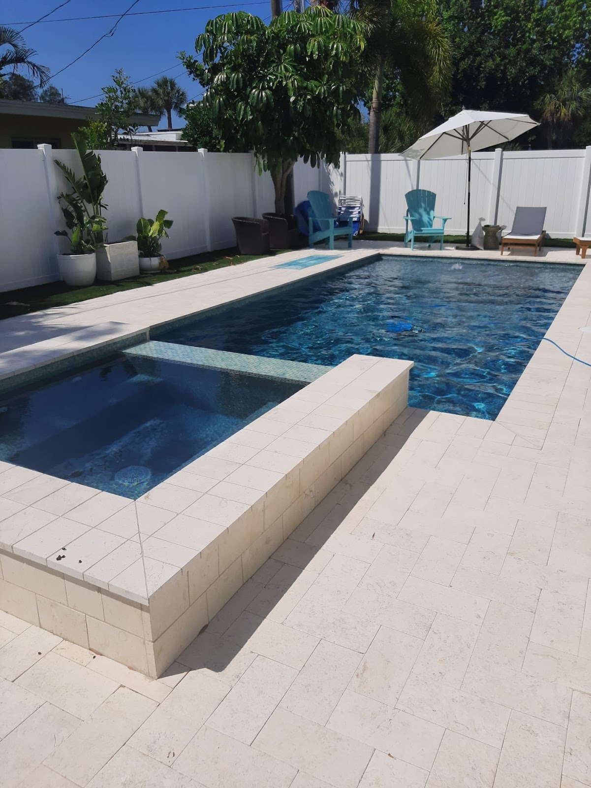 Modern rectangular backyard swimming pool with an attached raised spa, dark blue water, light paver decking, and a white privacy fence.