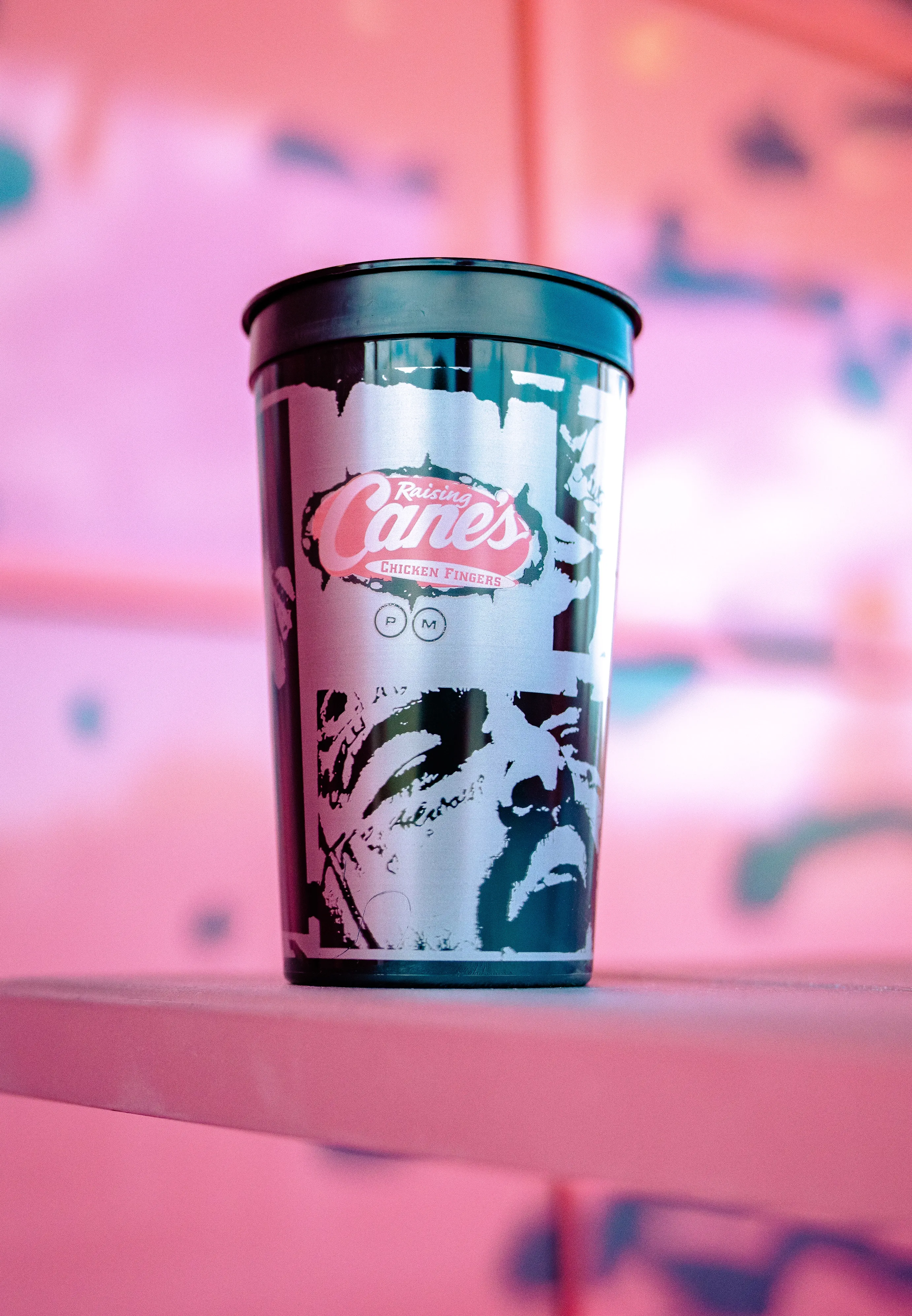 A close-up of a black souvenir cup sitting on a pink ledge. The cup features a silver, grunge-style graphic design depicting Post Malone, topped with the Raising Cane's logo in pink and a small "PM" circle icon. The background is a soft, out-of-focus pink wall.