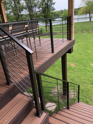 A custom elevated deck by Cedar Works Deck Builders features rich brown composite decking, black metal cable railings, and a matching stair system overlooking a spacious green backyard and water view. This finished outdoor living installation showcases the kind of modern, durable deck design Cedar Works Deck Builders creates for homeowners in Commerce Charter Twp, Michigan.