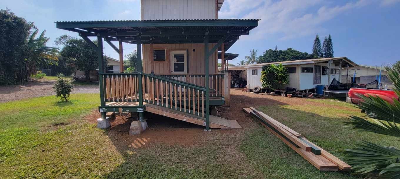 A small, beige, two-story structure with a dark green porch and covered deck. The porch features a wooden railing and a small ramp leading to a dirt path and grassy yard.