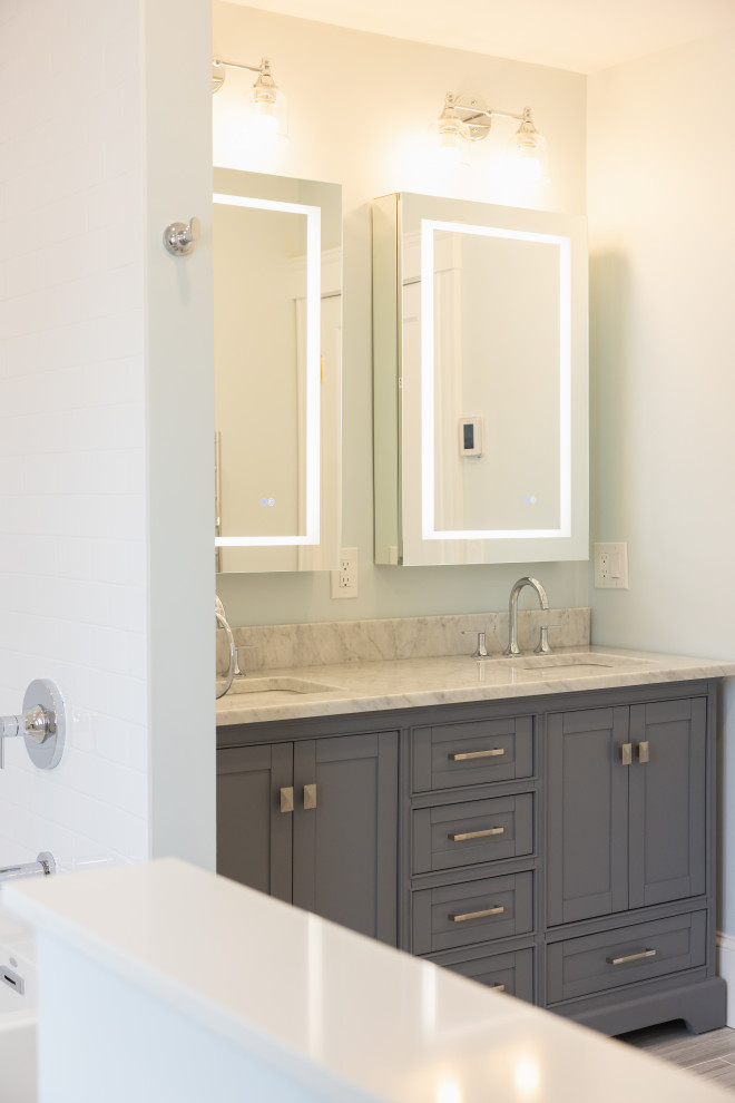 A modern bathroom vanity with a gray cabinetry, marble countertop, two sinks, and illuminated mirrors above. A chrome shower fixture is visible on the left. The overall aesthetic is clean and contemporary.
