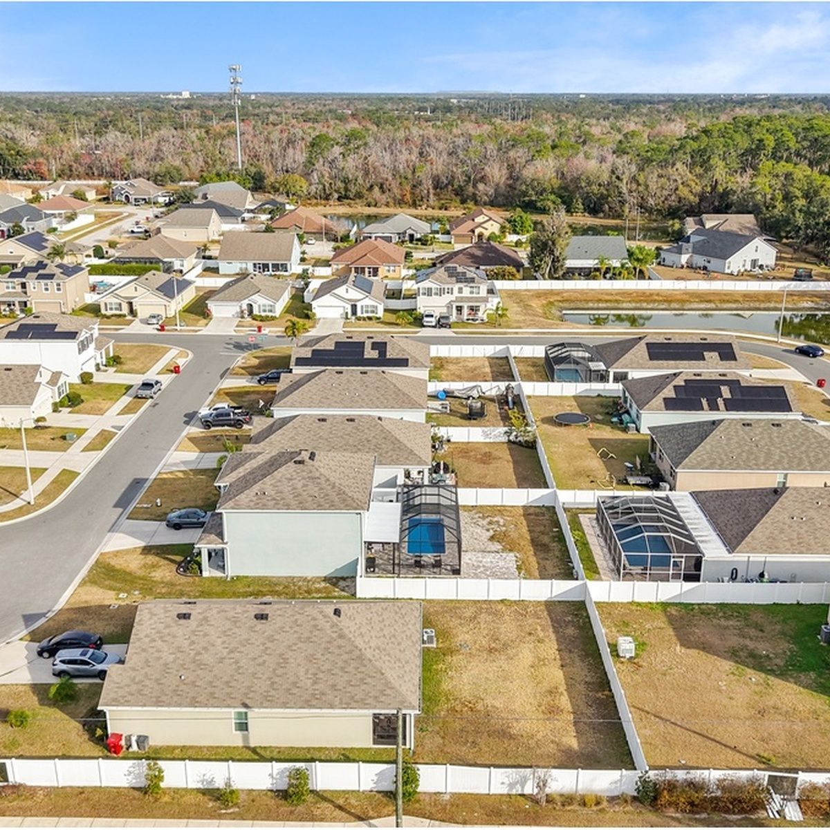 High-altitude drone shot of a quiet residential neighborhood featuring homes with brown and gray shingle roofs, enclosed pool lanai structures, and a retention pond nearby.