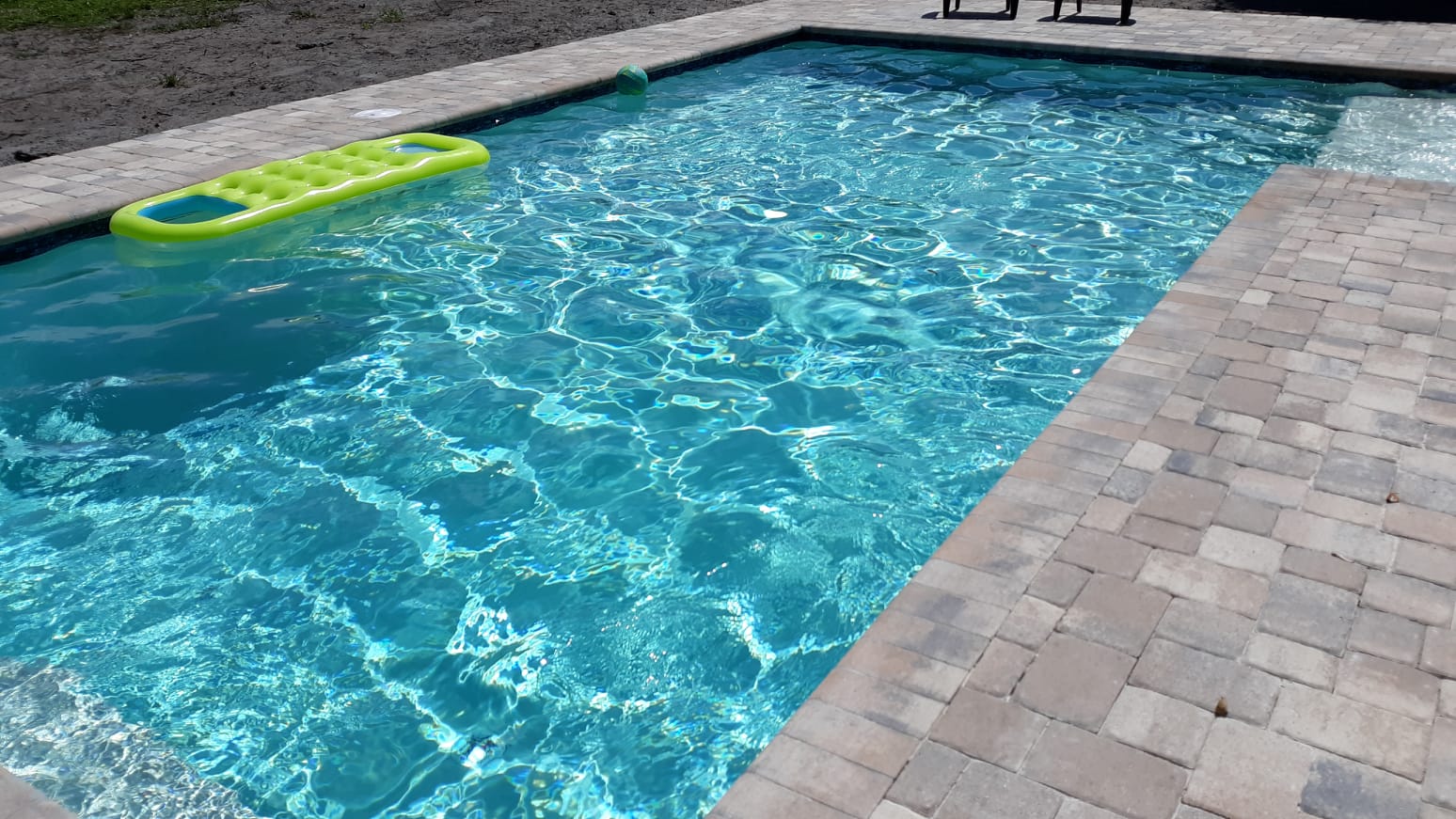Rectangular backyard swimming pool with sparkling blue water, surrounding brick pavers, and a pool float.
