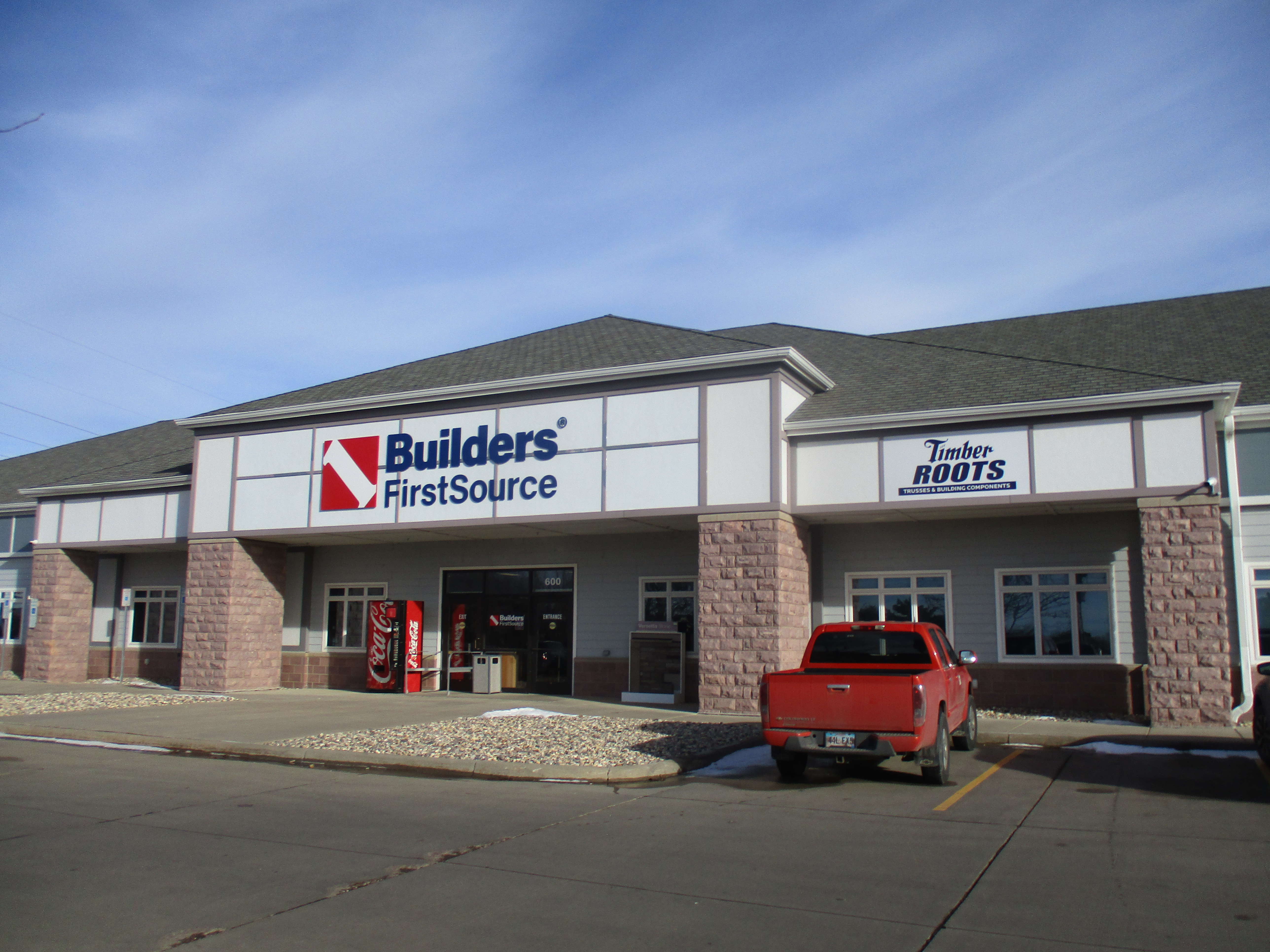 The exterior of the Builders FirstSource store, a modern building with a gray facade, white trim, and a bold Builders FirstSource sign prominently displayed. To the right is a sign for "Timber Roots Trusses & Building Components." A red pickup truck is parked in front of the building, and the sky is a bright blue with no clouds.