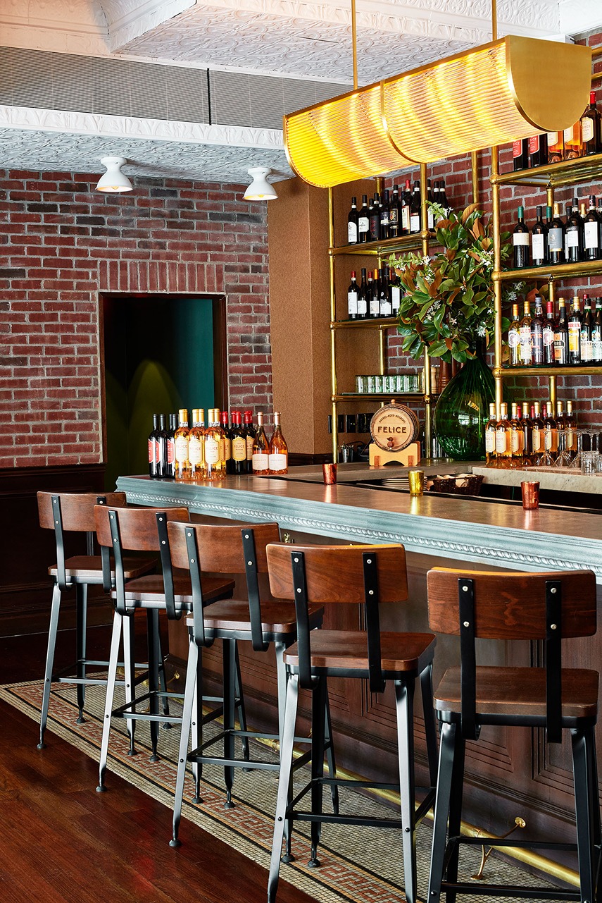Focused view of a dark wood bar with a metal trim and high-backed wooden stools. Bottles of ros&eacute; and red wine are staged on the counter under a glowing ribbed glass pendant light.