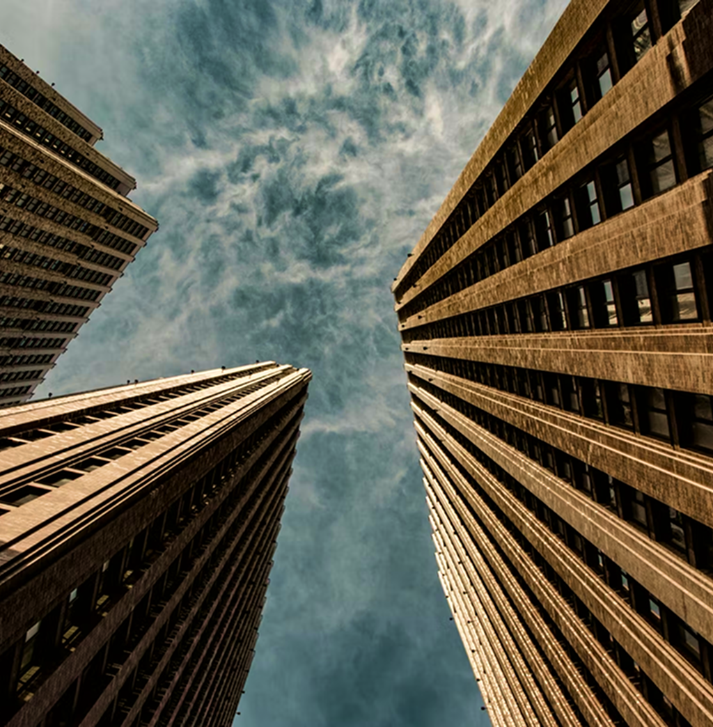 Upward view of three towering brutalist style office buildings against the backdrop of a partly cloudy sky.