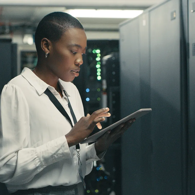 A Black woman in a white collared shirt and grey skirt is working on a tablet in a server room. She is wearing a lanyard with an ID badge. The server room has rows of server racks with blinking lights.