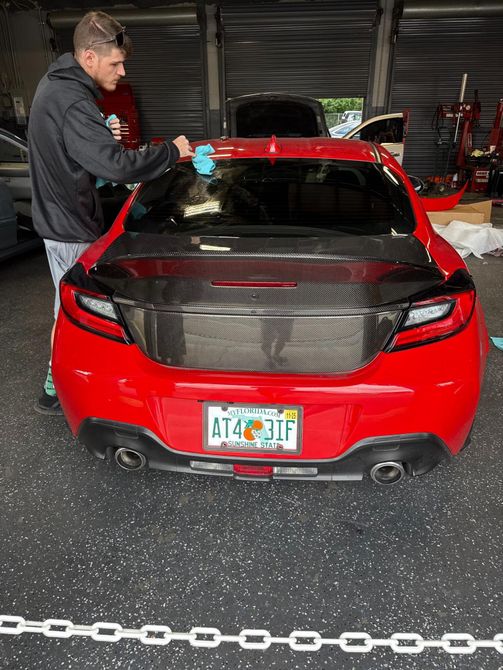 A technician is wiping down the dark-tinted rear window and carbon-fiber trunk lid of a shiny red sports car, likely a Subaru BRZ or Toyota 86, emphasizing detailing and aftermarket performance part installation.