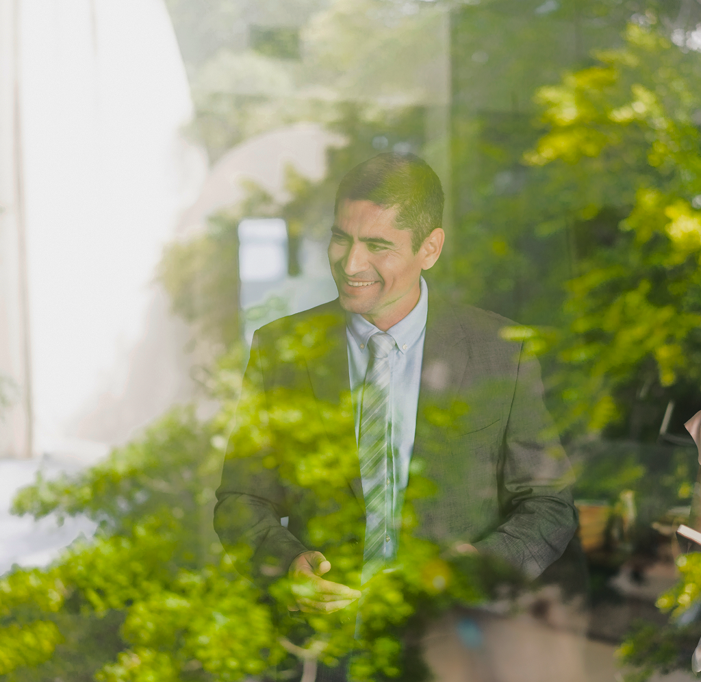 A suited professional standing in an office, as seen through a window with trees reflected in it.