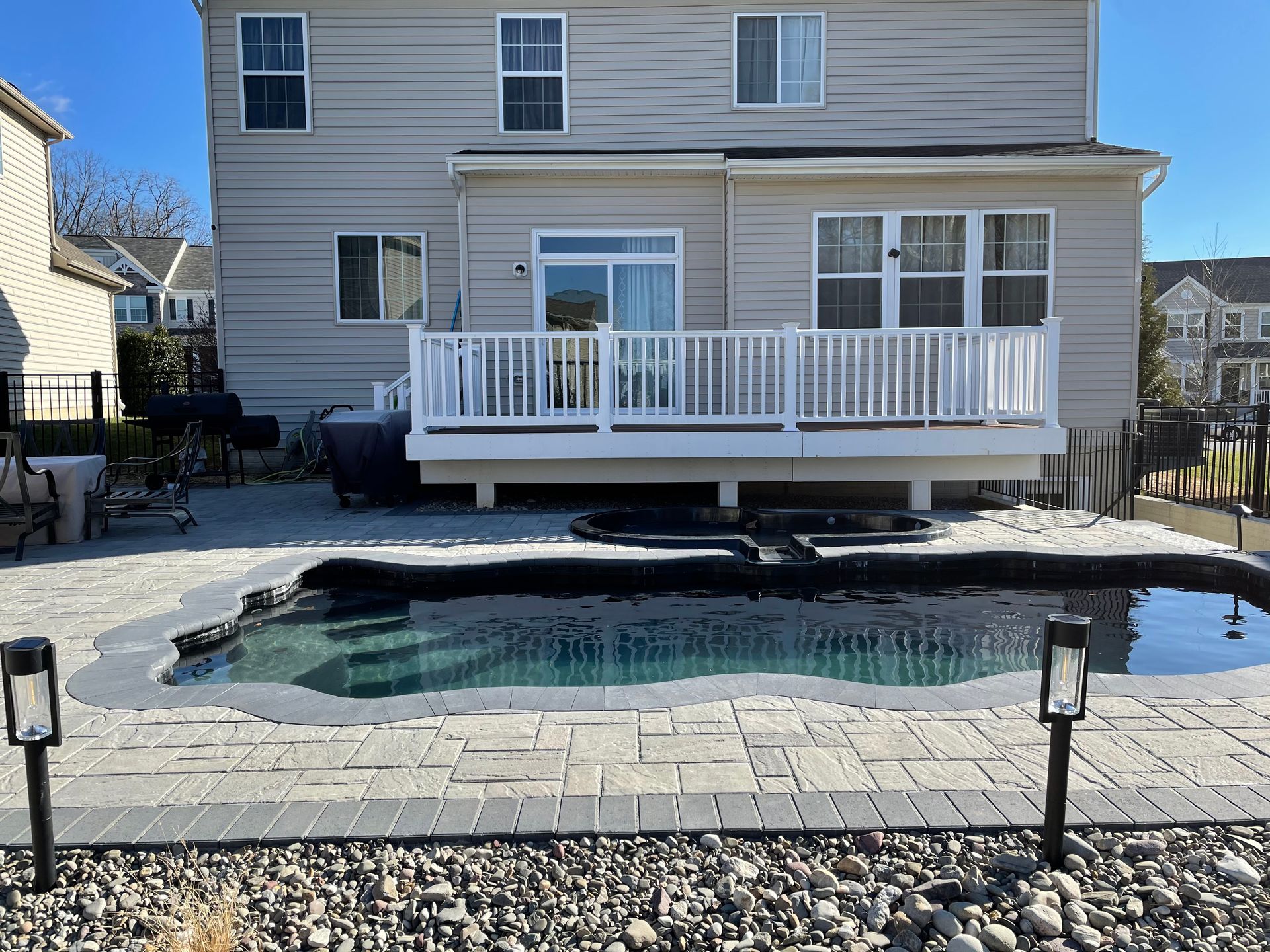 Front-facing view of backyard pool with paved patio, lighting fixtures, and landscaped stone border. The setup demonstrates complete outdoor design combining hardscaping, lighting, and functional layout for a polished residential environment.