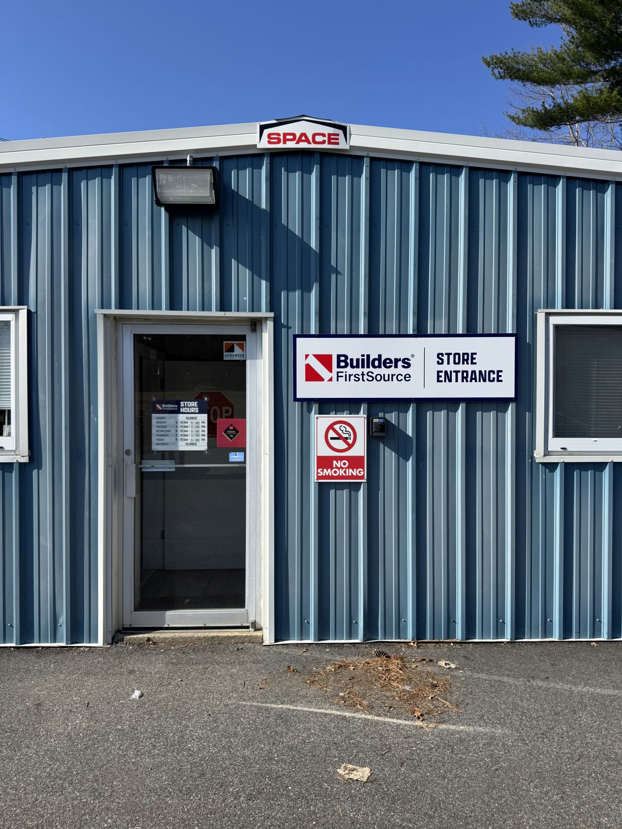 Builders FirstSource store entrance on a blue metal building, featuring a glass door, store entrance sign, and posted safety notices at a building materials and lumber supply location.