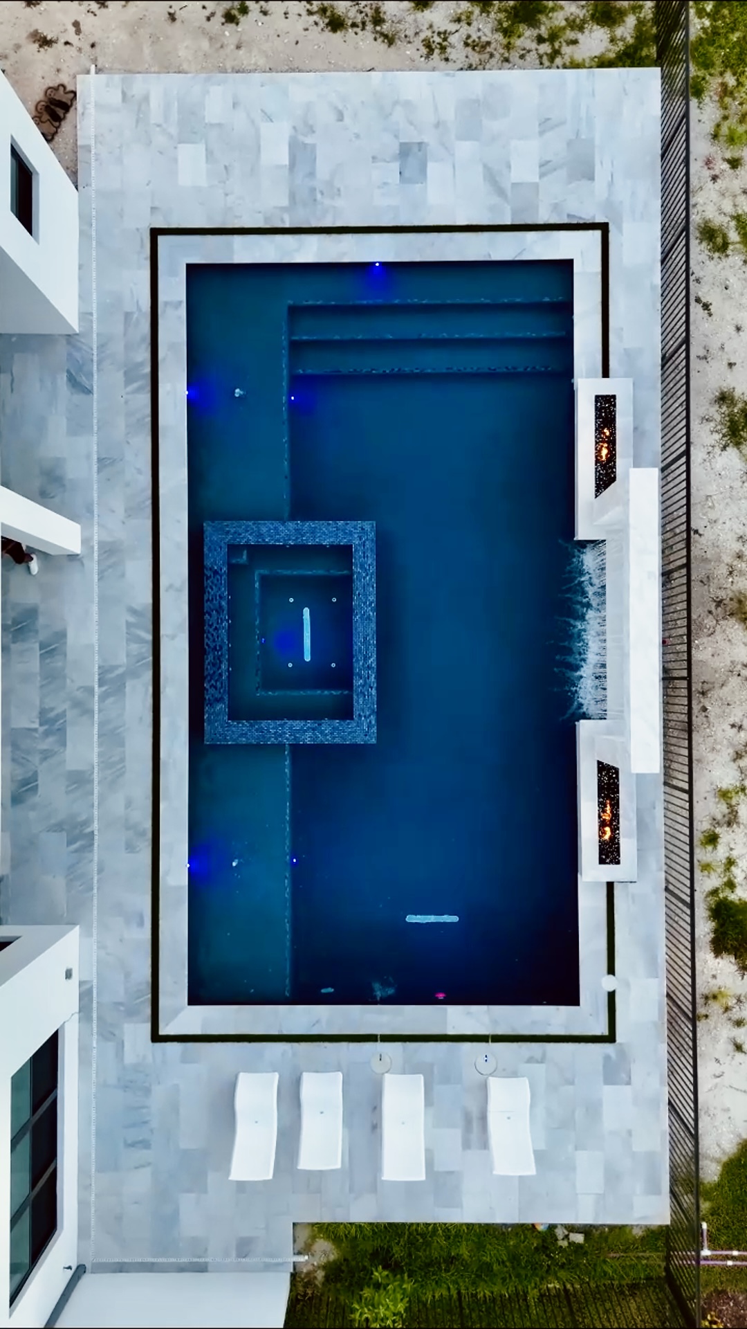 Top-down shot of a rectangular pool with spa, surrounded by a white stone patio and outdoor seating, emphasizing geometric design.