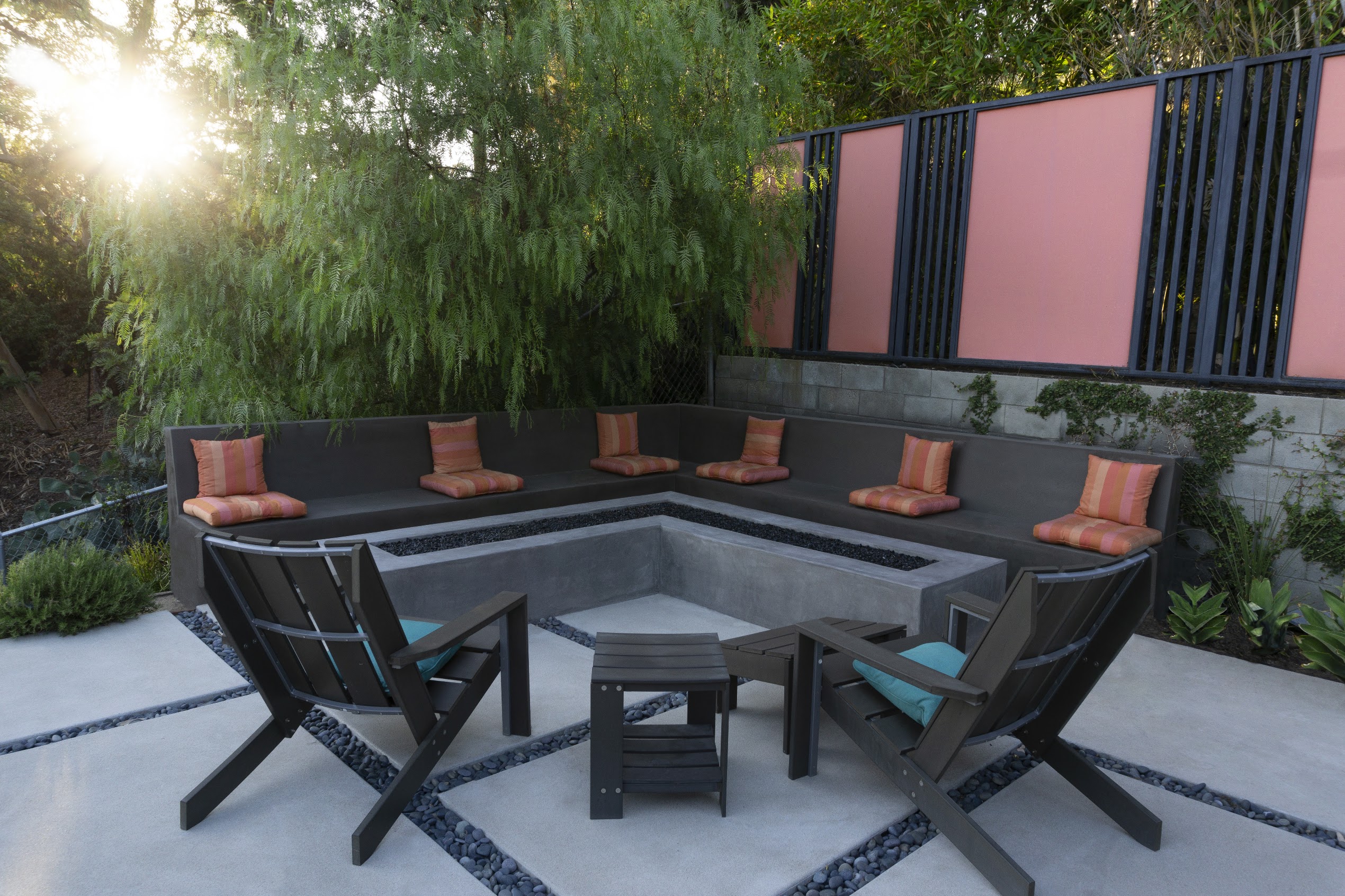 Modern sunken outdoor lounge with black seating, orange cushions, fire pit, and chairs, surrounded by greenery and warm evening sunlight