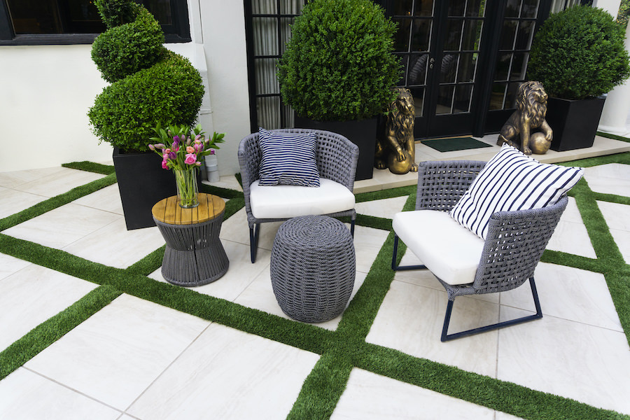 Modern outdoor patio with gray wicker chairs, striped pillows, flower vase, topiary plants, and geometric artificial grass accents.