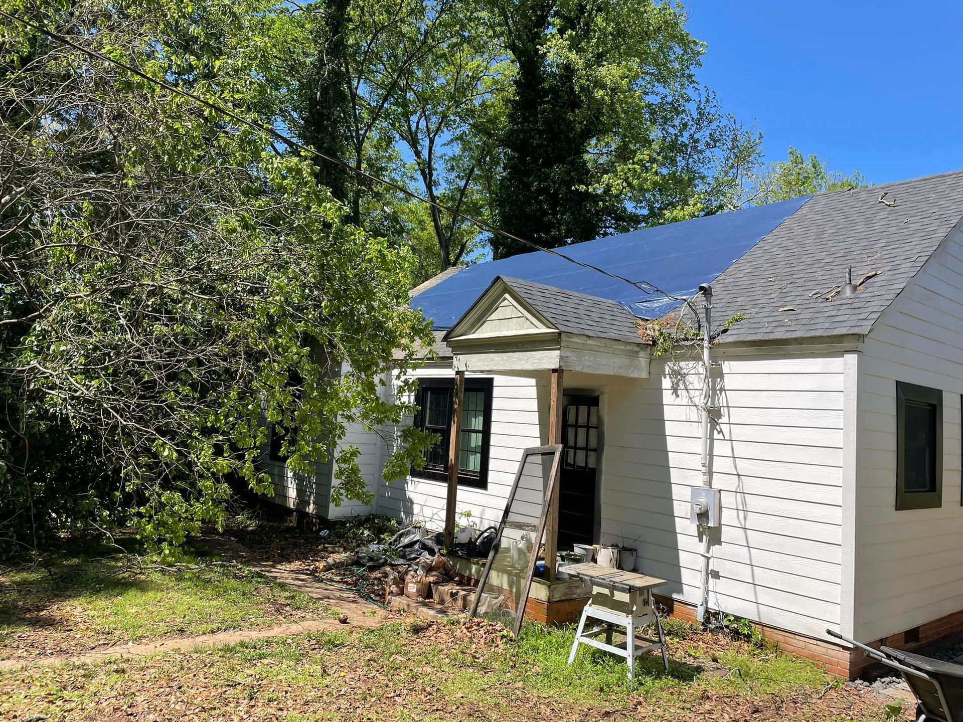 Residential house with solar panels on the roof surrounded by trees, showing yard clutter and maintenance work, including tools and debris near entrance, under clear blue sky in suburban setting.