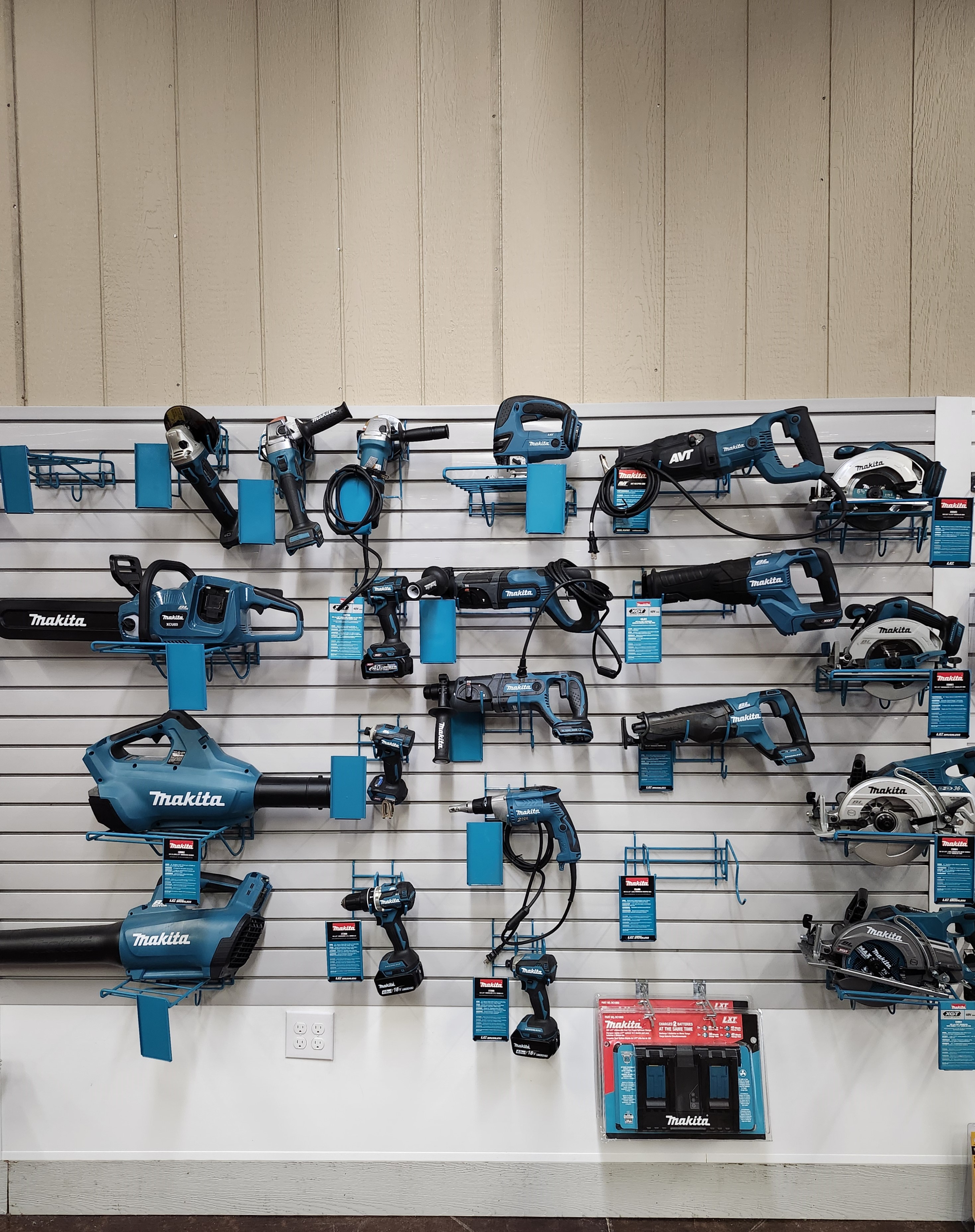 A well-organized display of Makita power tools and accessories at a Builders FirstSource lumber yard. The display features a range of drills, saws, blowers, and other power tools, all neatly hung on a slat wall with clear Makita branding and product information.