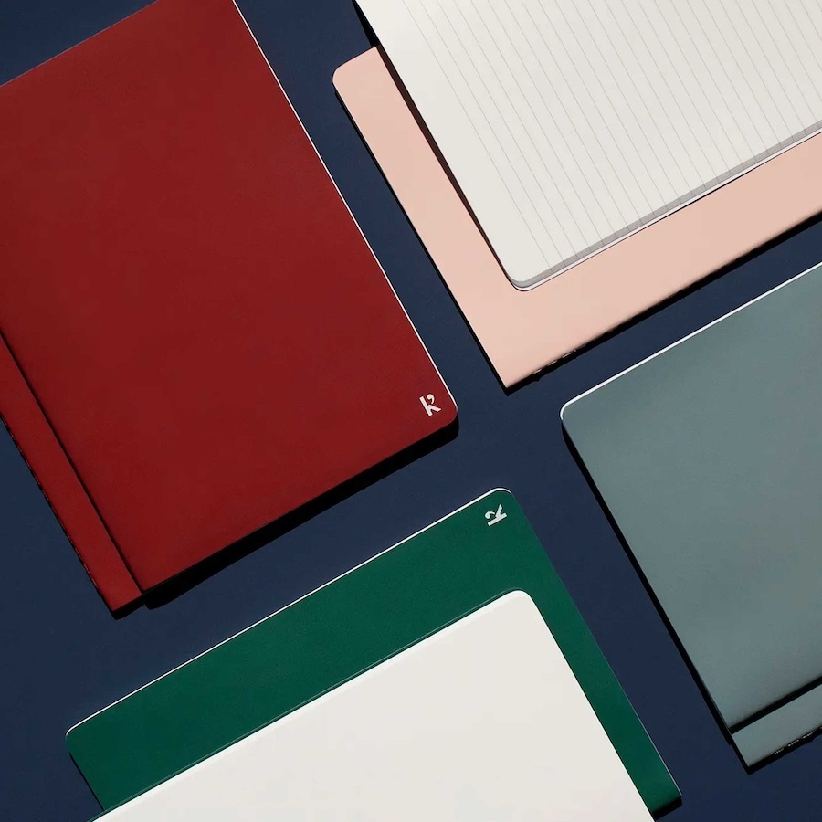 A dynamic overhead product image by Tabula Rasa Social Stationers, featuring a variety of Karst Stone Paper A5 journals. The display includes notebooks in vibrant red, sophisticated pink, deep green, and sleek grey, some open to reveal their signature smooth, lined stone paper. This visual highlights Karst's commitment to sustainability, presenting an innovative, tear-resistant, and waterproof alternative to traditional paper, crafted entirely from recycled stone for a refined writing experience.
