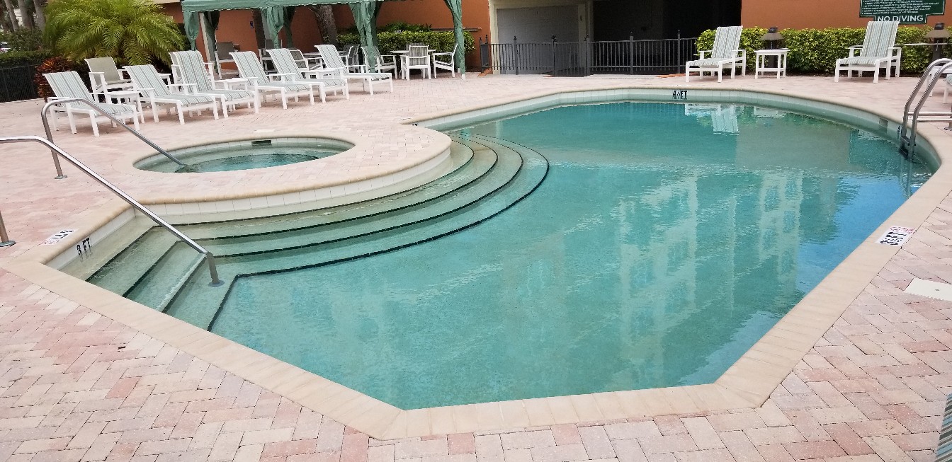 Community swimming pool with wide curved entry steps, light blue water, brick paver decking, and surrounding lounge seating.