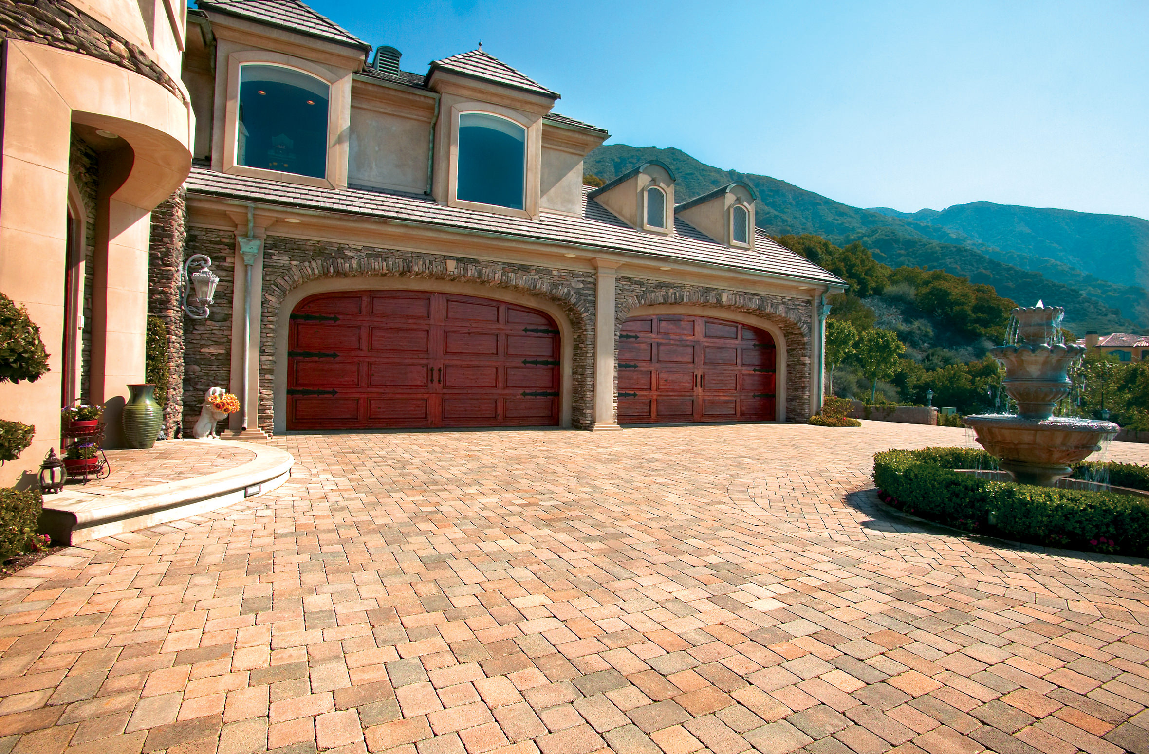 Luxury Mediterranean villa with twin red garage doors, brick-paved driveway, fountain, and mountain views beneath a clear blue sky.