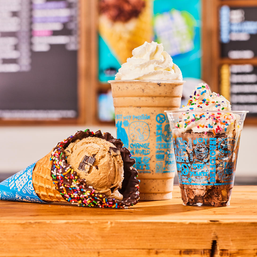 Waffle cone, shake and sundae in front of Ben & Jerry's ice cream shop menu board.